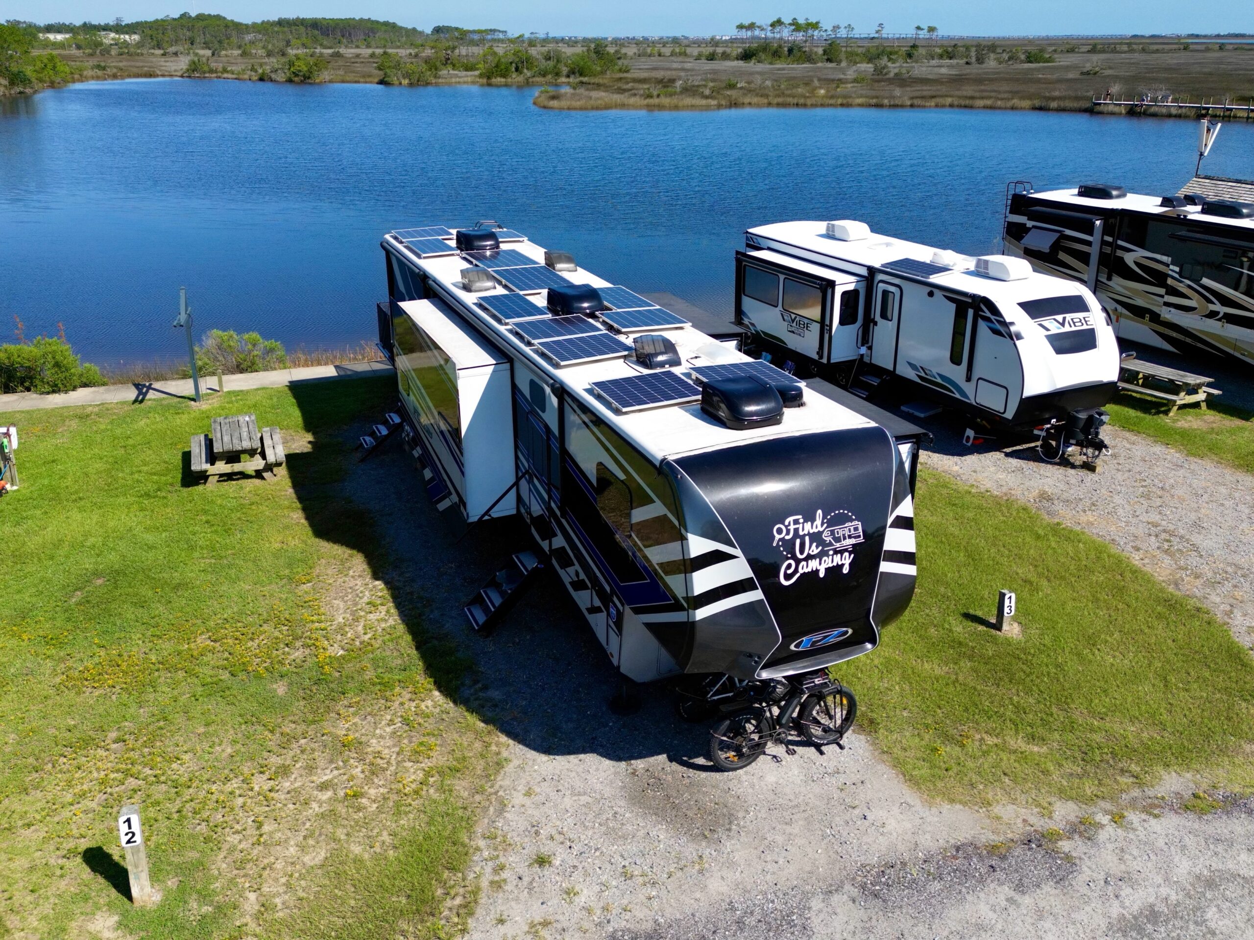 Aerial view of a Finduscamping RV with solar panels on top, backed up to the water. The RV is parked in a sunny spot, and the solar panels are absorbing the sunlight. The RV is surrounded by greenery, and the water is a beautiful blue color.